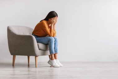 Genç Asyalı bir kadın yüzünü ellere gömer ve ağlar, depresyonda hisseder, beyaz stüdyo duvarına karşı koltukta oturur.