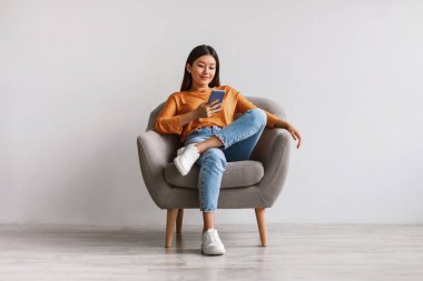 Akıllı telefon, internet ya da sosyal medya kullanan pozitif Asyalı kadın, koltukta oturuyor, yeni mobil uygulamaları kontrol ediyor