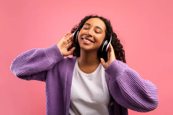 Радісна тисячолітня чорна жінка слухає музику з закритими очима, одягнена в навушники на рожевому студійному фоні — стокове фото