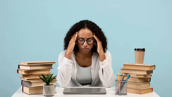 Studentessa nera esausta che studia a tavola con laptop e mucchi di libri su sfondo blu studio, panorama — Foto Stock