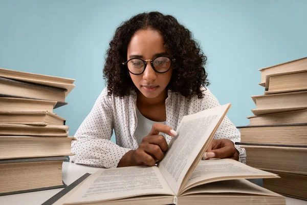 Estudiante negra enfocada sentada en el escritorio con montones de libros, leyendo, preparándose para el examen sobre fondo azul —  Fotos de Stock