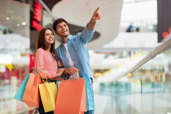 Szczęśliwa para Holding torby na zakupy wskazujące palcem stoi w centrum handlowym — Zdjęcie stockowe