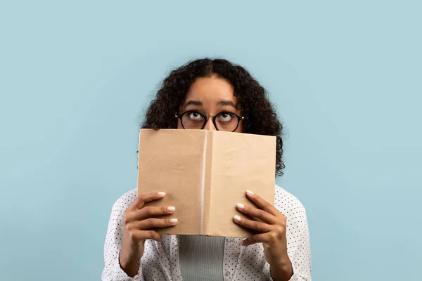 Gözlüklü havalı Afrikalı Amerikalı kız öğrenci kitabın arkasına saklanıyor, mavi stüdyo geçmişi üzerine üniversite sınavlarına çalışıyor. — Stok fotoğraf