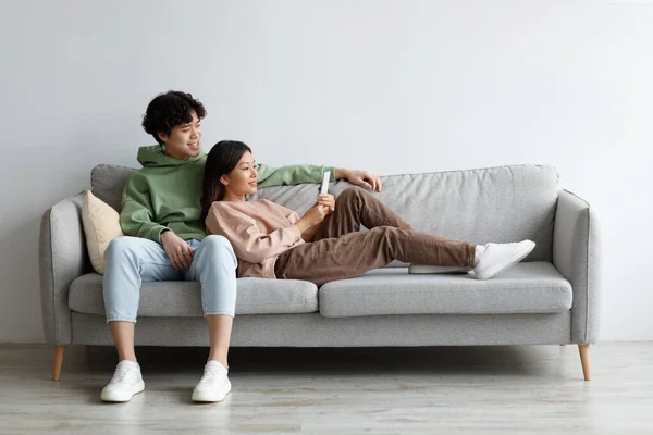 Jovem casal asiático afetuoso usando smartphone, relaxando no sofá juntos em casa, espaço livre — Fotografia de Stock