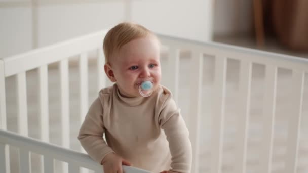 Adorable bébé avec sucette dans la bouche debout dans le berceau et agitant la main — Video