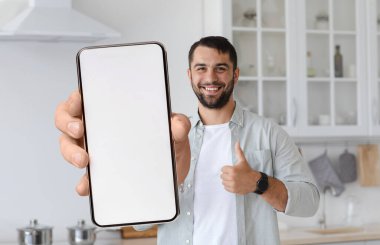 Çevrimiçi teklif. Neşeli Genç Adam Mutfakta Duruyor ve Boş Akıllı Telefonu Gösteriyor