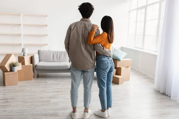 Πίσω όψη του χιλιετή άνθρωπος αγκαλιάζει τη σύζυγό του στο σαλόνι του νέου σπιτιού την ημέρα κίνησης, πλήρους διάρκειας. Έννοια μετεγκατάστασης — Φωτογραφία Αρχείου