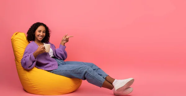 Millennial μαύρη γυναίκα πίνοντας καφέ, αναπαύεται σε καρέκλα τσάντα φασολιών, δείχνοντας κατά μέρος κενό χώρο σε ροζ φόντο — Φωτογραφία Αρχείου