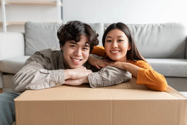 Portrait de heureux jeune couple asiatique penché sur une grande boîte en carton, posant et souriant à la caméra le jour en mouvement à l'intérieur — Photo