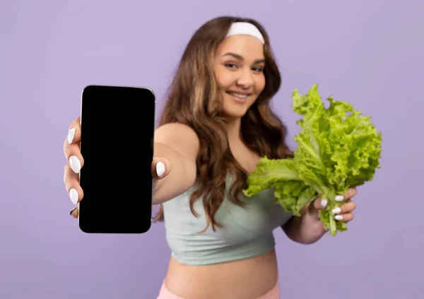 Sorridente caucasico plus size giovane atleta donna in uniforme sportiva mostra foglie di lattuga e smartphone con schermo bianco — Foto Stock