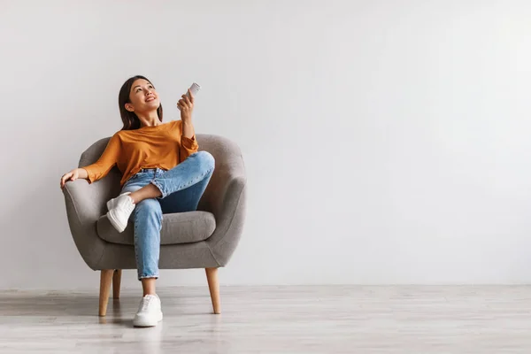 Ευτυχισμένη νεαρή Ασιάτισσα γυναίκα με τηλεχειριστήριο χαλαρώνοντας κάτω από το κλιματιστικό, κάθεται στην πολυθρόνα ενάντια στο λευκό τοίχο, mockup — Φωτογραφία Αρχείου