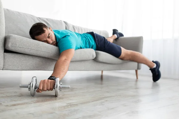 Fatigué mâle millénaire avec barbe se trouve sur le canapé avec haltères se reposant après l'exercice dans le salon intérieur — Photo