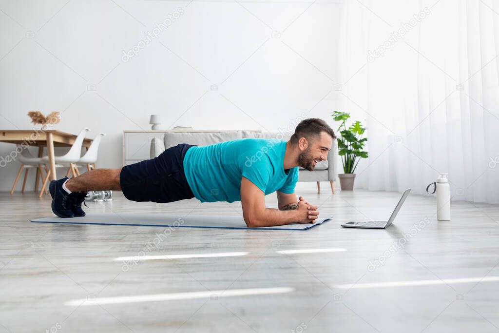 Happy young handsome guy athlete with beard do plank and watch fit lesson in living room