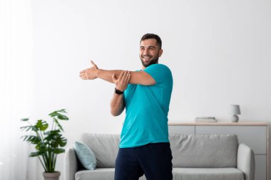 Sakallı neşeli genç erkek sporcu kollarını esnetir, kaslarını çalıştırır, vücudunu geliştirmeye çalışır.