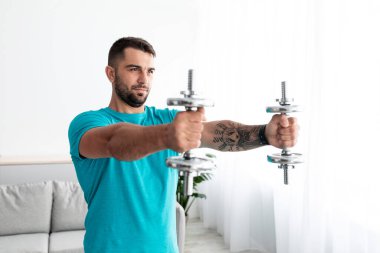 Mavi tişörtlü ciddi kaslı yakışıklı milenyum erkeği minimalist oturma odasında dambılları kaldırıyor.