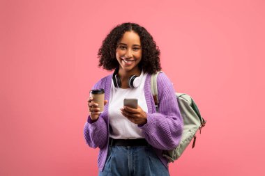 Sırt çantalı ve kulaklıklı mutlu siyah kadın hazır kahve içiyor, cep telefonu kullanıyor, internetten mesajlaşıyor.