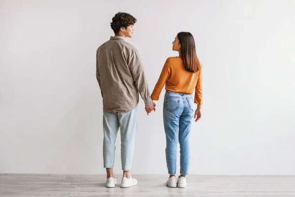Full längd av unga asiatiska man och kvinna som står med ryggen mot kameran, tittar på varandra och håller varandra i handen — Stockfoto