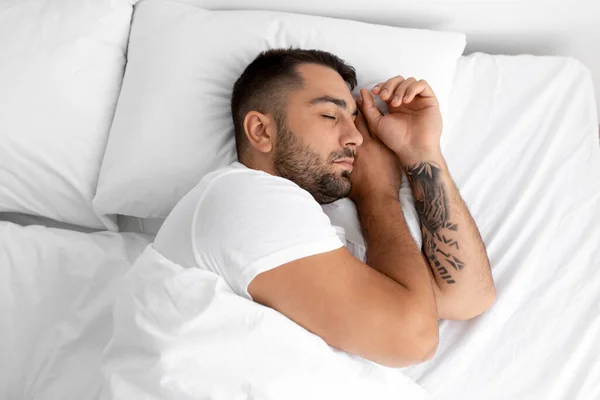 Спокойный миллениальный красавец с бородой спит на белой удобной кровати на мягкой подушке в спальне, вид сверху — стоковое фото