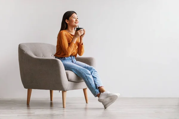 Vacker ung asiatisk kvinna dricker kaffe eller te, har paus, avkopplande i fåtölj mot vit studio vägg — Stockfoto