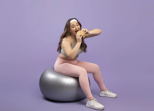 Gelukkig grappig vrij hongerig Kaukasisch plus size jonge dame in sportuniform zit op fit bal en het eten van hamburger — Stockfoto