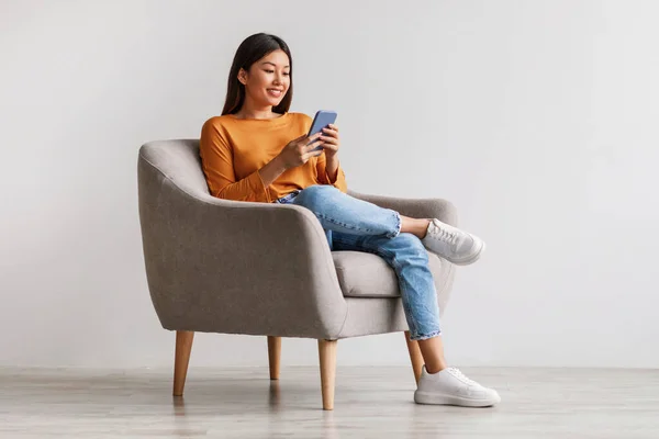 Mujer asiática encantadora usando el teléfono celular, comunicándose en la web, trabajando o aprendiendo en línea, sentado en sillón, espacio libre —  Fotos de Stock