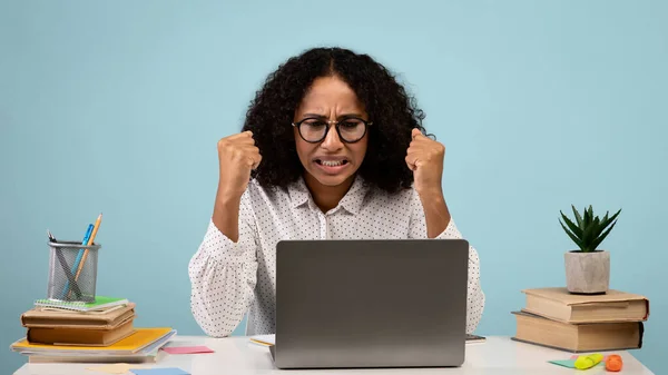 Ung svart kvinna i glasögon sitter vid skrivbordet, tittar på bärbar skärm, knyta näven, känna sig arg på blå bakgrund — Stockfoto