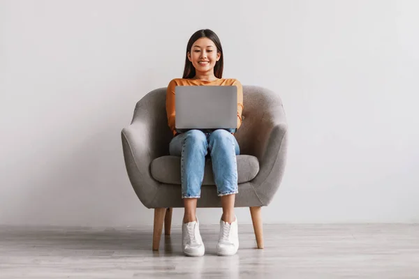 Όμορφη νεαρή Ασιάτισσα που κάθεται στην πολυθρόνα, δουλεύοντας σε λάπτοπ ενάντια σε λευκό τοίχο στούντιο, πλήρους μήκους — Φωτογραφία Αρχείου