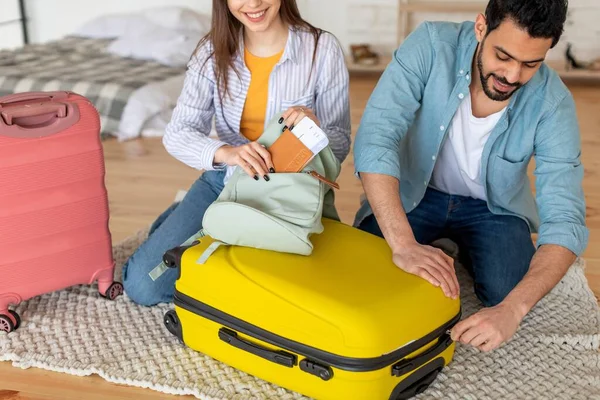 Jóvenes cónyuges empacando ropa en maleta y mujer poniendo pasaporte con boletos en bolsa, preparándose para las vacaciones — Foto de Stock