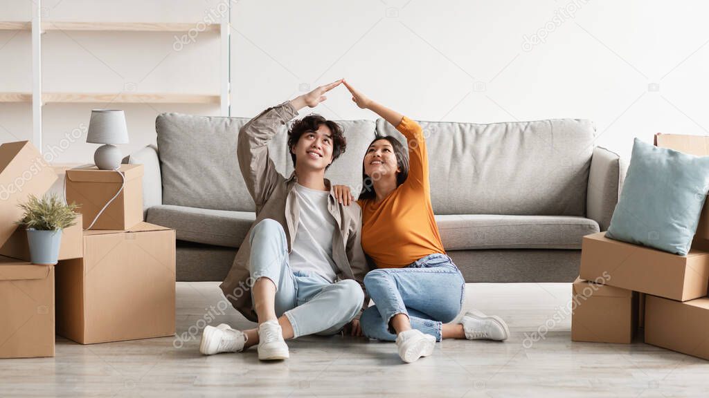 Cheery millennial Asian couple relocating to new home, making house roof gesture, sitting on floor among carton boxes