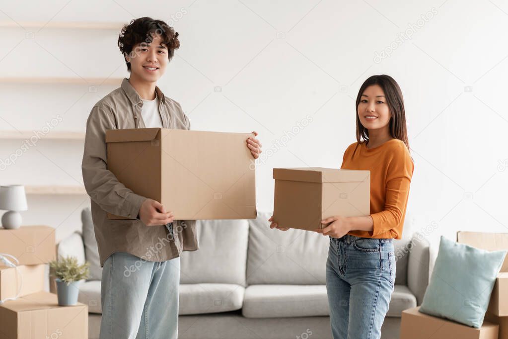 Cheerful young Asian woman and her husband holding carton boxes, smiling at camera in new home. Moving day concept