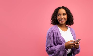 Akıllı telefon tutan güzel Afrikalı genç bir bayan, pembe stüdyo arka planında boş bir alana bakıyor.