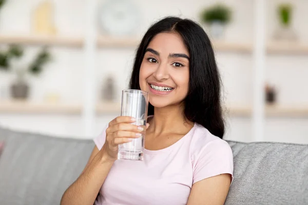 Tersenyum wanita muda India memegang gelas air bersih segar sambil duduk di sofa di ruang tamu. Jauhkan konsep terhidrasi — Stok Foto