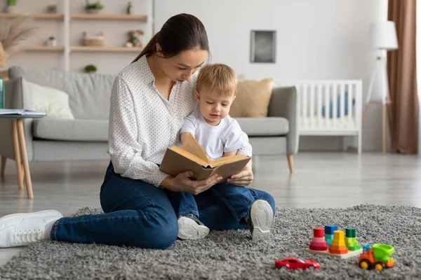 Bebek gelişimi. Genç anne, küçük oğluyla evde kitap okuyor, anne çocuğuyla vakit geçiriyor. — Stok fotoğraf