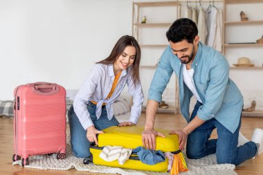 Elbise dolu bavulu kapatmaya çalışan genç çift, tatile hazırlanıyor ve evde birlikte bir şeyler hazırlıyorlar.