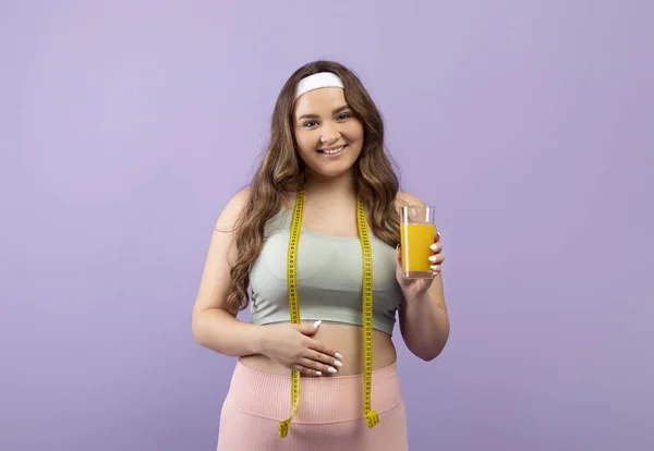 Happy pretty caucasian plus size young woman in sportswear with measuring tape show glass of fresh orange juice — Stock Photo, Image