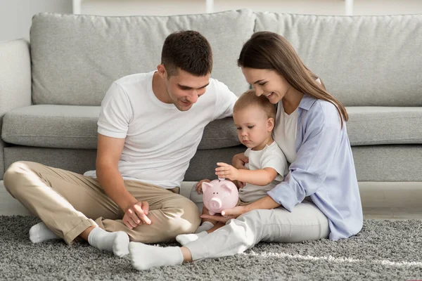 Presupuesto familiar. Padres felices mirando al hijo pequeño poniendo monedas a Piggybank —  Fotos de Stock