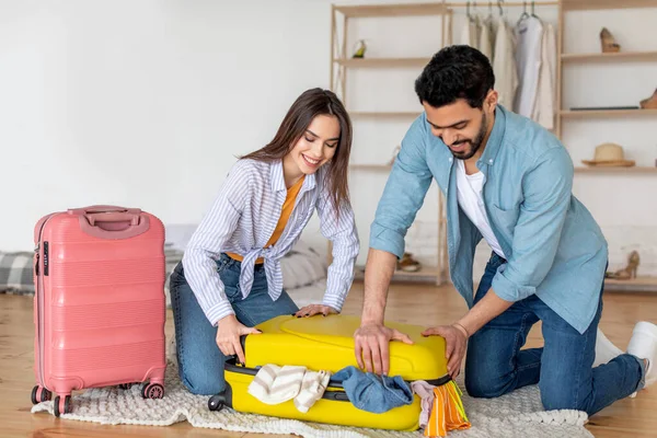 Pareja joven tratando de cerrar la maleta llena de ropa, preparándose para las vacaciones y empacando cosas en casa — Foto de Stock