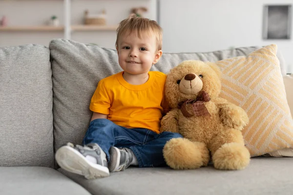 かわいいです小さな幼児男の子座っているソファの近くテディベアと見てカメラ,かわいいです子供遊びとともにおもちゃ — ストック写真