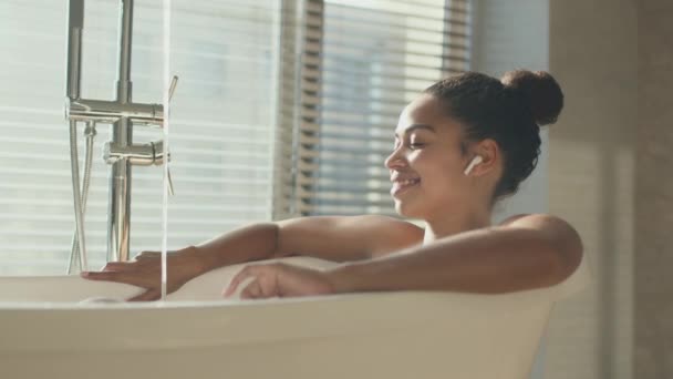 Jeune femme afro-américaine profitant de la musique dans des écouteurs sans fil, se reposant dans un bain à remous avec de l'eau coulante, zoom avant — Video
