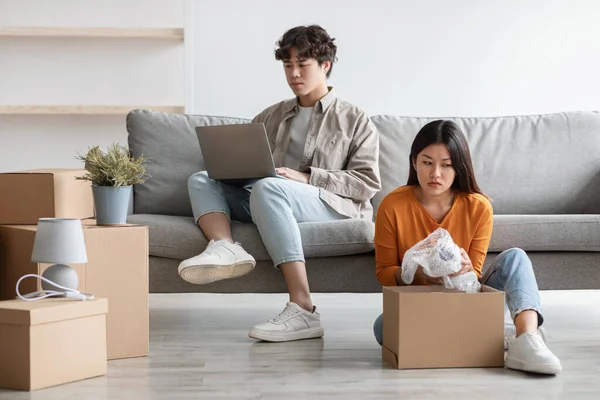Joven mujer asiática desembalaje caja de cartón en el día de mudanza en un nuevo hogar, mientras que su novio usando el ordenador portátil, jugando videojuegos —  Fotos de Stock