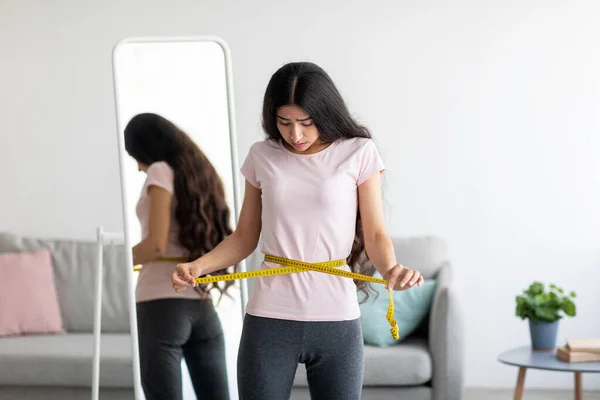 Unzufriedene junge Inderin misst Taille mit Klebeband in der Nähe des Spiegels und ärgert sich über Ergebnisse der Gewichtsabnahme-Diät zu Hause — Stockfoto