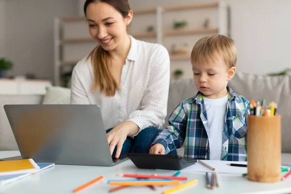 Freiberufliches und Mutterschaftskonzept. Junge berufstätige Mutter mit Laptop und ihr kleiner Junge spielen mit Taschenrechner in der Nähe der Mutter — Stockfoto