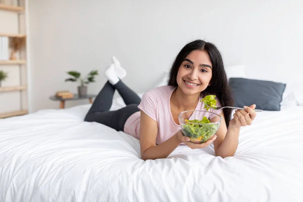 Piękna młoda Hinduska kobieta jedząca sałatkę warzywną leżąc na łóżku w domu, wolne miejsce. Zrównoważona dieta surowcowa — Zdjęcie stockowe