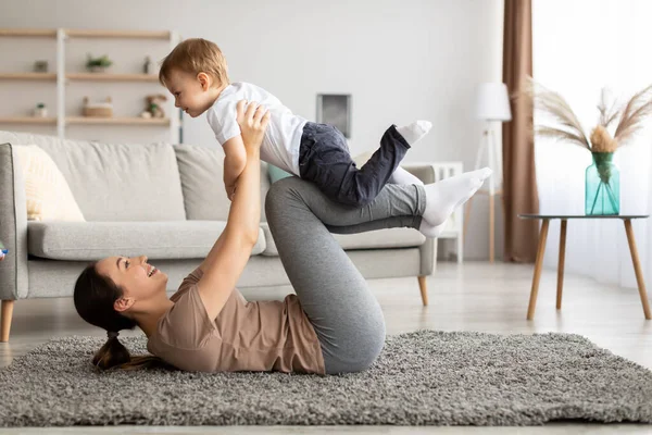 Jonge moeder traint samen met haar peuter zoon, balanceert kind op benen, doet fitness training in de woonkamer — Stockfoto