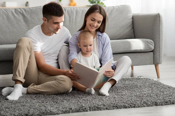 Família feliz de três com o filho infantil leitura livro juntos em casa — Fotografia de Stock