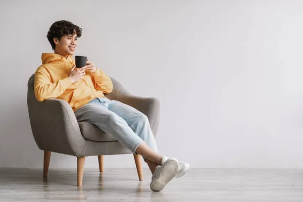 Longitud completa de hombre asiático joven bebiendo café caliente en sillón contra la pared del estudio blanco, espacio vacío —  Fotos de Stock