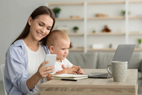 Zdalna kariera. Młoda mama z dzieckiem na rękach przy użyciu smartfona i laptopa — Zdjęcie stockowe