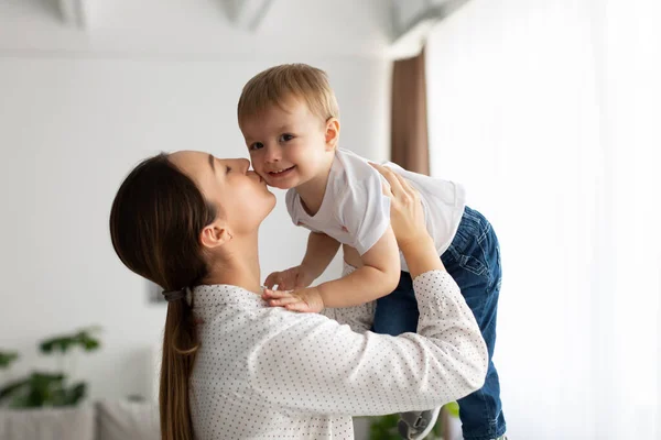 Concepto de infancia feliz. Joven madre caucásica besando a su pequeño hijo en la mejilla, sosteniéndolo en el aire y jugando en casa —  Fotos de Stock