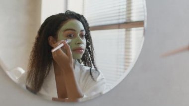 Genç Afrikalı Amerikalı kadın organik kilden bir maske takıyor, banyoya bakıyor, boş alan arıyor.