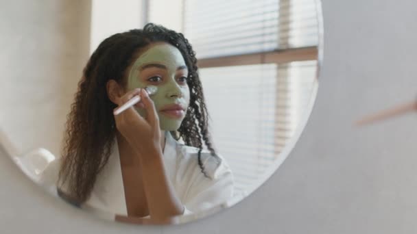 Mujer afroamericana joven aplicando mascarilla de arcilla orgánica nutritiva en la cara, mirando el espejo en el baño, espacio libre — Vídeo de stock